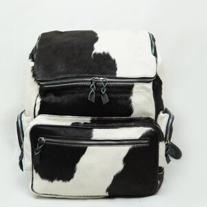 May include: A black and white cowhide backpack with two zippered compartments. The backpack has a top flap with a zipper closure and adjustable straps.
