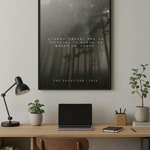 Può includere: Un poster incorniciato con una scena di foresta nebbiosa e il testo "QUANDO PREGHI PER LA PIOGGIA. IL FANGO VA MESSO IN CONTO" e "THE EQUALIZER 2014". Il poster è appeso a una parete bianca sopra una scrivania con un laptop, una lampada e una pianta in vaso.