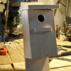 Peut inclure: Un nichoir en bois avec un trou d'entrée circulaire et un petit perchoir. Le nichoir est fabriqué en bois de couleur claire et a une forme rectangulaire. Il est placé sur un fond d'atelier avec des outils et des machines.