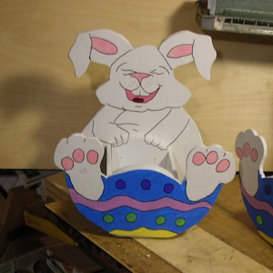 Peut inclure: Un lapin en bois blanc assis dans un œuf de Pâques bleu, jaune, rose et vert. Le lapin sourit et a des oreilles et des pieds roses.