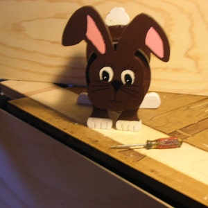 Peut inclure: Une figurine de lapin en bois brun avec des accents peints en blanc, y compris de grands yeux, une queue moelleuse et des pieds. Le lapin est debout sur une surface en bois avec un tournevis rouge et argenté à côté.