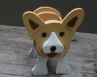 May include: A wooden corgi dog planter painted in shades of tan, white, and brown. The planter has a smiling face with a white snout, brown ears, and a red tongue. The dog is standing on four white legs.