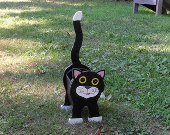 May include: A black cat-shaped planter with a white-tipped tail and white paws stands on green grass. The cat has large yellow eyes, pink ears, and a smiling face. The planter is made of wood and is designed to hold plants.