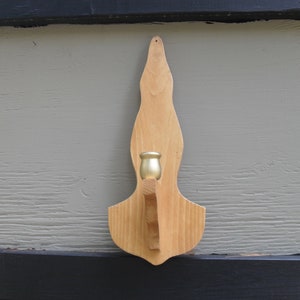 Peut inclure: Un bougeoir mural en bois avec une finition naturelle. Il présente une forme décorative et une coupelle de couleur laiton pour accueillir une bougie. Ce support est conçu pour être accroché au mur et ajoute une touche rustique à la décoration intérieure.