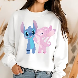 Pu&ograve; includere: Una felpa bianca a maniche lunghe con una stampa grafica del personaggio blu Stitch e del personaggio rosa Angel. I due personaggi sono in piedi l'uno accanto all'altro, con Angel che si sporge per baciare Stitch sulla guancia.