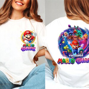 May include: Off-white t-shirt featuring a colorful graphic of Mario from the Super Mario Galaxy video game. The front design shows Mario waving, with the words "Mario Galaxy". The back of the shirt displays a larger, detailed scene with multiple characters and the text "The Super Mario Galaxy".