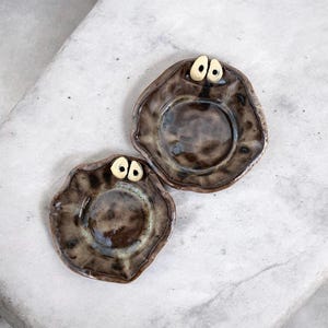 Könnte beinhalten: Zwei handgefertigte Keramikschalen mit einem einzigartigen Design. Die Schalen sind braun mit glänzendem Finish und weisen ein skurriles, augenähnliches Detail auf. Die unregelmäßige Form und die Farbvariationen verleihen ihnen einen rustikalen, künstlerischen Reiz. Diese dekorativen Schalen eignen sich perfekt zum Servieren kleiner Portionen.