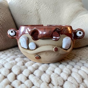 Könnte beinhalten: Eine handgefertigte Keramik-Garnschale mit einem monsterartigen Gesicht, bestehend aus einem glänzend rotbraunen Oberteil und einem matten beigen Unterteil. Die Schale hat zwei hervorstehende Augen an den Seiten, weiße Zähne und eine Öffnung.