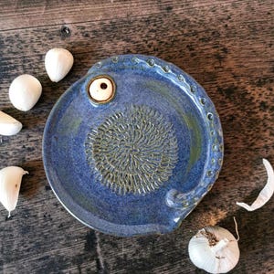 Könnte beinhalten: Ein runder, blauer Knoblauchreibe-Teller aus Keramik mit einer strukturierten, sternförmigen Mitte zum Reiben und einem kleinen, erhabenen weißen Keramikknopf am Rand. Der Teller weist eine Reihe kleiner, kreisförmiger Vertiefungen am Rand auf.