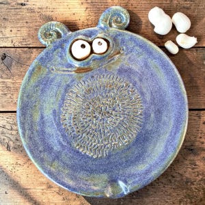 Könnte beinhalten: Ein runder, violetter und grüner Keramik-Knoblauchreibe-Teller mit einem stilisierten Monstergesicht, zwei spiralförmigen Ohren, großen weißen Augen und einer strukturierten Oberfläche in der Mitte zum Reiben. Drei weiße Knoblauchzehen liegen daneben.