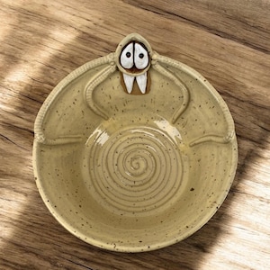 Könnte beinhalten: Eine gesprenkelte, hellbraune Keramikschale mit einem skurrilen Monsterdesign. Die Schale hat ein Spiralmuster in der Mitte und ein Wesen mit großen Augen und Fangzähnen oben. Die Schale hat zwei kleine Griffe an den Seiten.