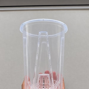 Könnte beinhalten: Ein transparenter, zylindrischer Pflanztopf aus Kunststoff mit einem erhöhten zentralen Entwässerungskegel und vertikalen Belüftungsschlitzen an den Seiten. Der Behälter hat einen breiten, flachen Rand oben und einen strukturierten Boden mit Löchern.