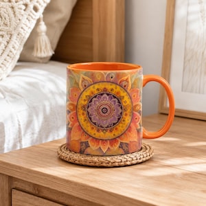 May include: A ceramic mug with a vibrant orange handle and interior, featuring a detailed, multi-layered mandala design in shades of yellow, orange, purple, and red. The mug rests on a round, woven natural fiber coaster.