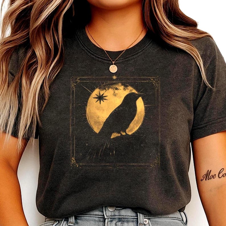 May include: Dark gray t-shirt featuring a gold illustration of a raven perched in front of a full moon and star. The design is framed within a gold rectangular border. The shirt has short sleeves and a crew neck.