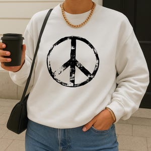 May include: White sweatshirt featuring a large, distressed black peace sign graphic. The long-sleeved top is paired with blue jeans. A black shoulder bag and a gold chain necklace and bracelet complete the look. A black coffee cup is held in the hand.