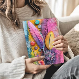 May include: A colorful journal with a textured cover featuring a large pink and purple feather, a yellow feather, and various round embellishments. The background has a mix of blue, yellow, and pink hues, creating a vibrant artistic design.