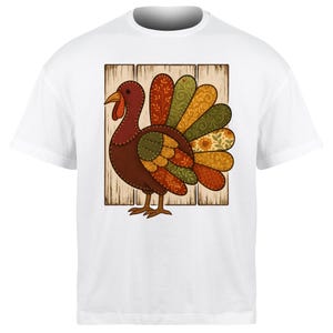 Puede incluir: Camiseta blanca con un diseño de pavo colorido. El pavo está hecho de varias telas estampadas en tonos marrones, rojos, naranjas y verdes, sobre un fondo de madera. El diseño está centrado en la camiseta, lo que la convierte en una prenda festiva.