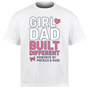 Pode incluir: Camiseta branca com o texto "GIRL DAD BUILT DIFFERENT" em letras pretas e rosa. O design inclui um coração e um laço rosa, com a frase "POWERED BY PIGTAILS & SASS" abaixo.