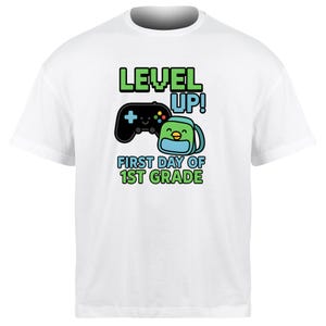 Op de afbeelding: Wit T-shirt met een grafisch ontwerp met de woorden "LEVEL UP!" in groen en blauw, een zwarte gamecontroller en een rugzak met een lachend gezicht. Daaronder staat "FIRST DAY OF 1ST GRADE."