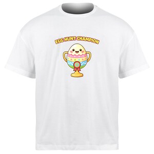 May include: White t-shirt featuring a cartoon trophy with an egg design. The egg has a smiling face and is decorated with colorful patterns. The text "EGG HUNT CHAMPION" is above the trophy in yellow.