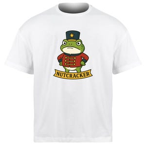 May include: White t-shirt featuring a cartoon frog dressed as a nutcracker. The frog is green with a red jacket and a black hat with a gold star. The word "NUTCRACKER" is printed on a yellow banner below the frog.