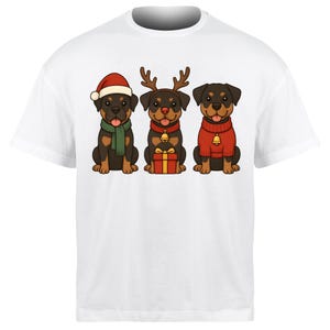 Puede incluir: Camiseta blanca con tres cachorros de Rottweiler de dibujos animados. Uno lleva un gorro de Papá Noel y una bufanda, otro cuernos de reno y un regalo, y el tercero un suéter rojo con una campana.