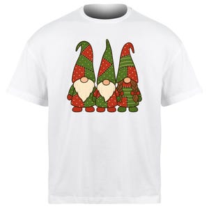 Könnte beinhalten: Weißes T-Shirt mit einem skurrilen Design von drei Wichteln. Die Wichtel haben rot-grüne Patchwork-Hüte und -Mäntel, cremefarbene Bärte und lächelnde Gesichter. Ein festliches Design für die Weihnachtszeit.