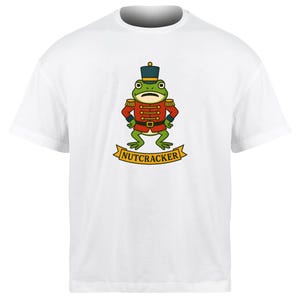 May include: White t-shirt featuring a cartoon frog dressed as a nutcracker. The frog is green with a red jacket, gold epaulettes, and a dark blue hat. The word "NUTCRACKER" is printed on a yellow banner below the frog.
