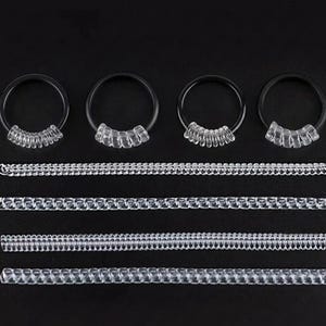 Puede incluir: Una colección de retenedores de piercing de plástico transparente y joyería de piercing de plástico transparente. Los retenedores son circulares con una serie de pequeñas cuentas transparentes. La joyería es recta y también tiene pequeñas cuentas transparentes.