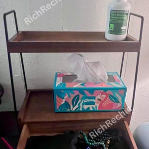 May include: A two-tiered wooden shelf with black metal frame. The top shelf holds a white bottle of headache relief. The middle shelf has a tissue box with a tropical design. The bottom drawer is open.