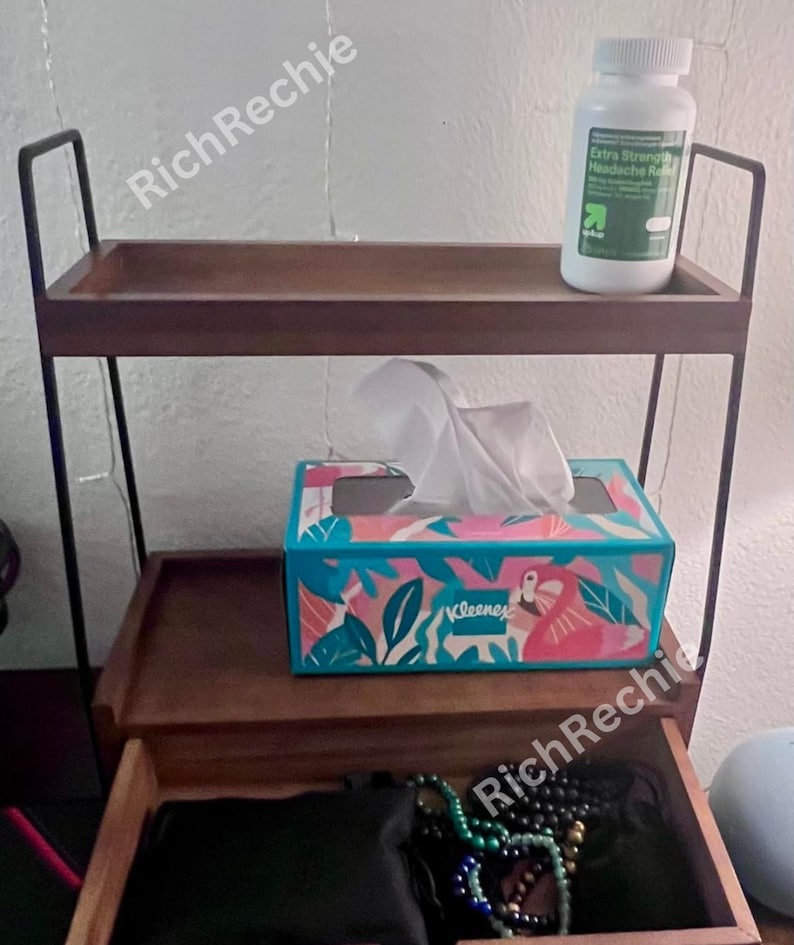 May include: A two-tiered wooden shelf with black metal frame. The top shelf holds a white bottle of headache relief. The middle shelf has a tissue box with a tropical design. The bottom drawer is open.