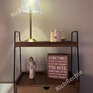 May include: A two-tiered wooden nightstand with a drawer, black metal frame, and a small lamp with a clear, ribbed shade. A candle in a glass jar and a decorative angel figurine are on the shelves. A wooden sign reads "Sometimes talking to your sister is all the therapy you need."