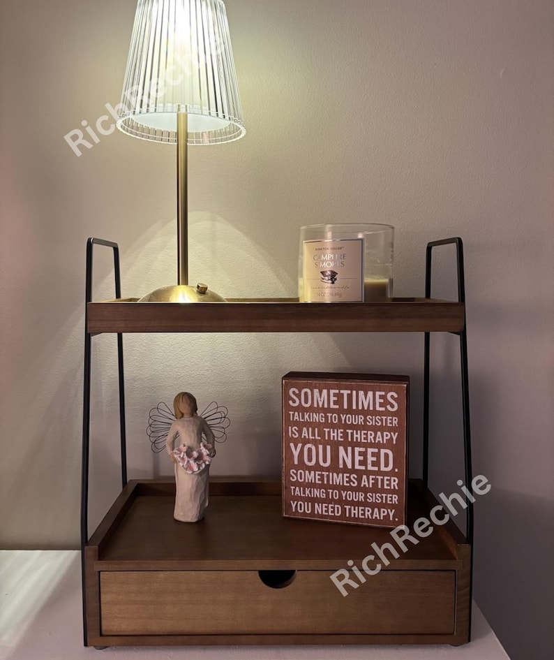 May include: A two-tiered wooden nightstand with a drawer, black metal frame, and a small lamp with a clear, ribbed shade. A candle in a glass jar and a decorative angel figurine are on the shelves. A wooden sign reads "Sometimes talking to your sister is all the therapy you need."