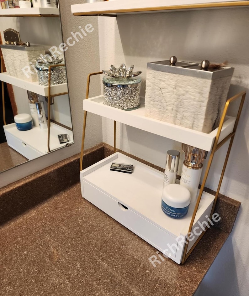 May include: A white and gold two-tiered shelf unit with a drawer, holding skincare products and decorative containers. The shelf has a modern design with gold metal supports and white shelves. The items on the shelves include a jar with a crystal lid and a textured box.