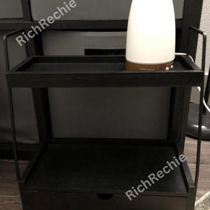 May include: A black two-tiered side table with a drawer. A white and brown essential oil diffuser sits on the top shelf. The table has a minimalist design with a rectangular top and bottom shelf and a drawer.