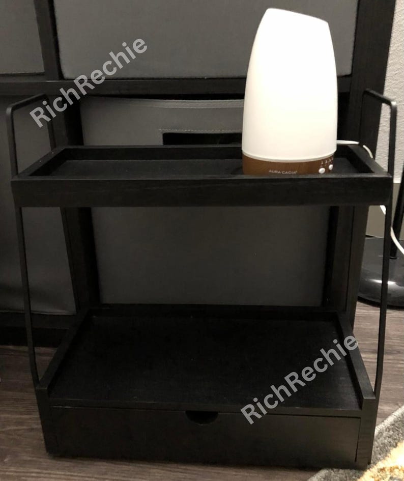 May include: A black two-tiered side table with a drawer. A white and brown essential oil diffuser sits on the top shelf. The table has a minimalist design with a rectangular top and bottom shelf and a drawer.