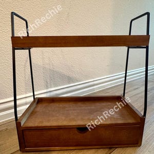 May include: A two-tiered wooden shelf with a drawer. The shelf has a dark brown finish and is supported by black metal frames. The top shelf has a raised edge, and the drawer has a round handle. The shelf is designed for storage and display.