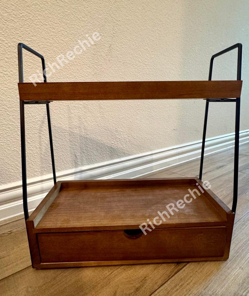 May include: A two-tiered wooden shelf with a drawer. The shelf has a dark brown finish and is supported by black metal frames. The top shelf has a raised edge, and the drawer has a round handle. The shelf is designed for storage and display.
