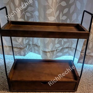 May include: A two-tiered, dark brown wooden shelf with a drawer. The shelf has a rectangular shape with a black metal frame. The top shelf is open, while the bottom shelf has a drawer with a small, round handle.