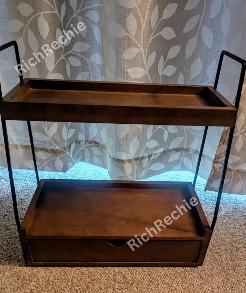 May include: A two-tiered, dark brown wooden shelf with a drawer. The shelf has a rectangular shape with a black metal frame. The top shelf is open, while the bottom shelf has a drawer with a small, round handle.