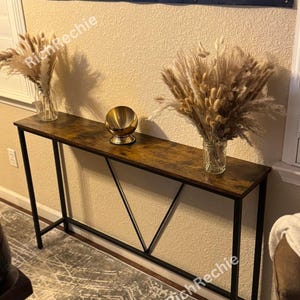 May include: A long, narrow console table with a dark brown wood top and black metal frame. The table features a decorative triangular support and is adorned with dried floral arrangements in glass vases and a gold sphere. A world map hangs on the wall.