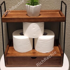May include: A two-tiered wooden bathroom shelf with a drawer, holding three rolls of white toilet paper and a small potted succulent. The shelf has black metal supports and is designed to sit atop a toilet.