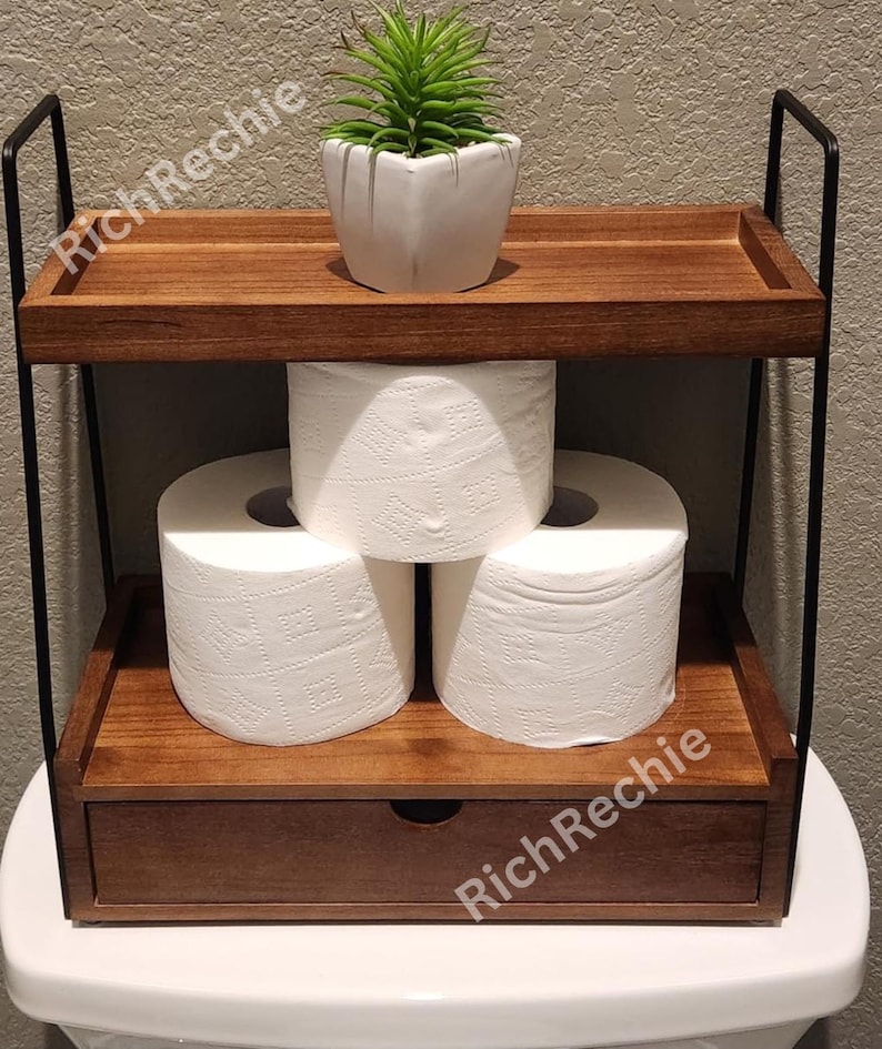 May include: A two-tiered wooden bathroom shelf with a drawer, holding three rolls of white toilet paper and a small potted succulent. The shelf has black metal supports and is designed to sit atop a toilet.