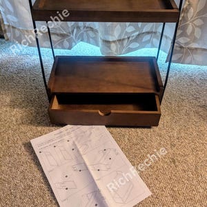 May include: A dark brown, three-tiered shelf with a drawer, supported by a black metal frame. The top two shelves are open, while the bottom shelf has a pull-out drawer. Assembly instructions are visible on the floor.