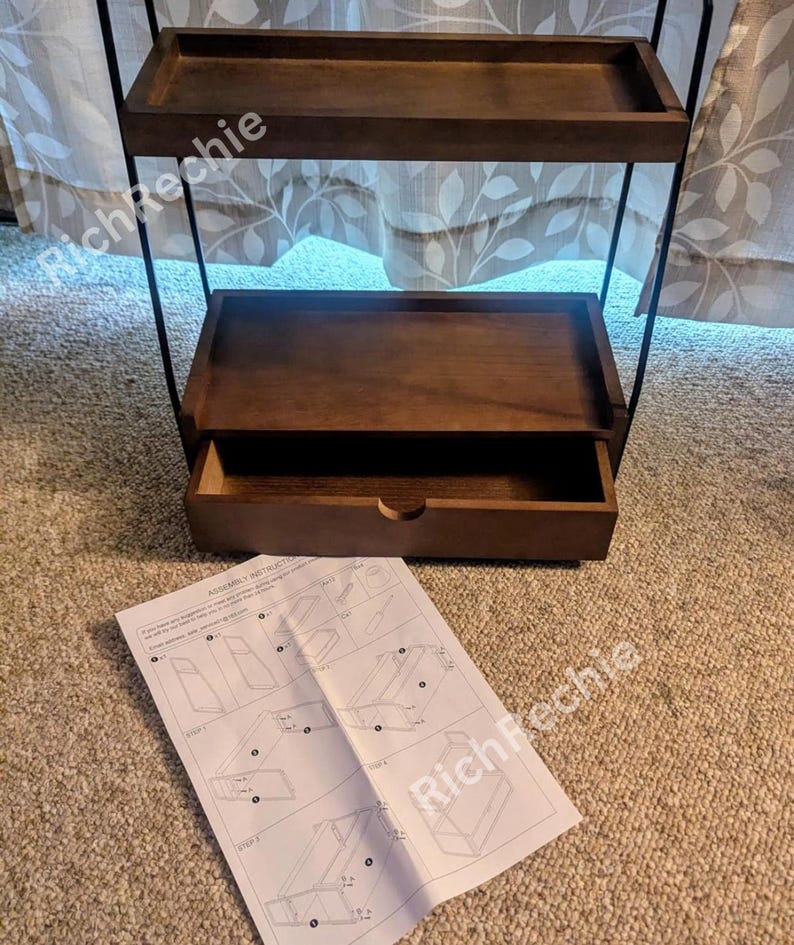 May include: A dark brown, three-tiered shelf with a drawer, supported by a black metal frame. The top two shelves are open, while the bottom shelf has a pull-out drawer. Assembly instructions are visible on the floor.