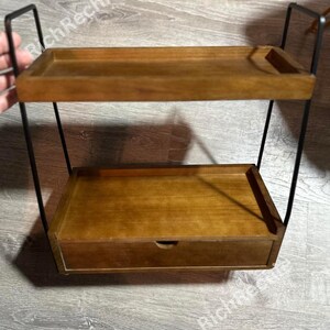 May include: Two-tiered wooden shelf with a drawer, supported by a black metal frame. The shelves and drawer are a warm brown color. The top shelf has a small raised edge on one side. The drawer has a small, round handle.