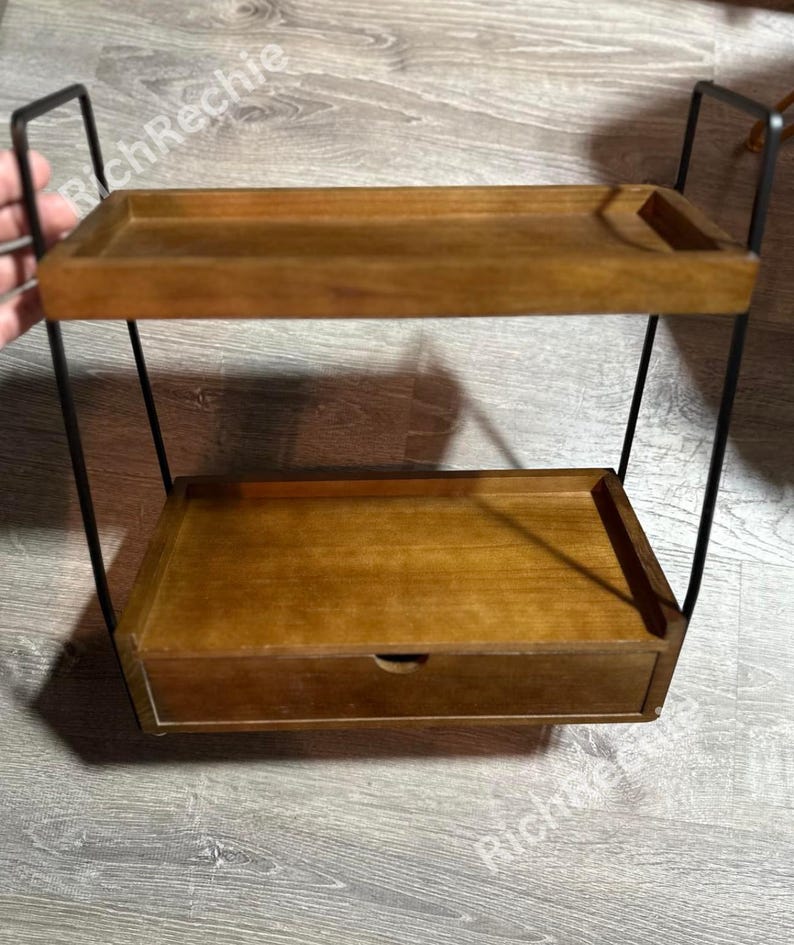 May include: Two-tiered wooden shelf with a drawer, supported by a black metal frame. The shelves and drawer are a warm brown color. The top shelf has a small raised edge on one side. The drawer has a small, round handle.