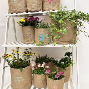 Könnte beinhalten: Ein weißes, mehrstöckiges Regal zeigt verschiedene Jute-Pflanzgefäße, gefüllt mit bunten Blumen und Grünpflanzen. Einige Pflanzgefäße sind mit Stickereien wie einer Biene, einer Blume und den Texten "Happy New Home" und "World's Best Mum" verziert.