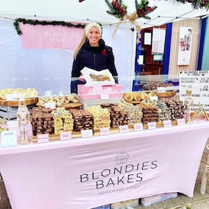 Op de afbeelding: Een roze marktkraam met een verscheidenheid aan gebak. De tafel is bedekt met een roze tafelkleed met de tekst "BLONDIES BAKES HOMEMADE CAKES". Een vrouw staat achter de tafel, die vol ligt met brownies en andere lekkernijen.
