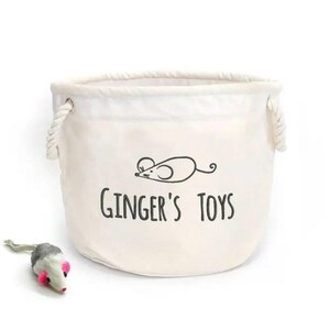 May include: A white canvas storage bin with rope handles and a black and white illustration of a mouse with the text "Ginger's Toys". A small grey and white toy mouse is in the foreground.